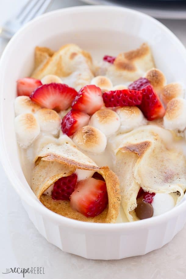 crepes rolled with strawberries marshmallows and chocolate in a white baking dish topped with marshmallows and strawberries