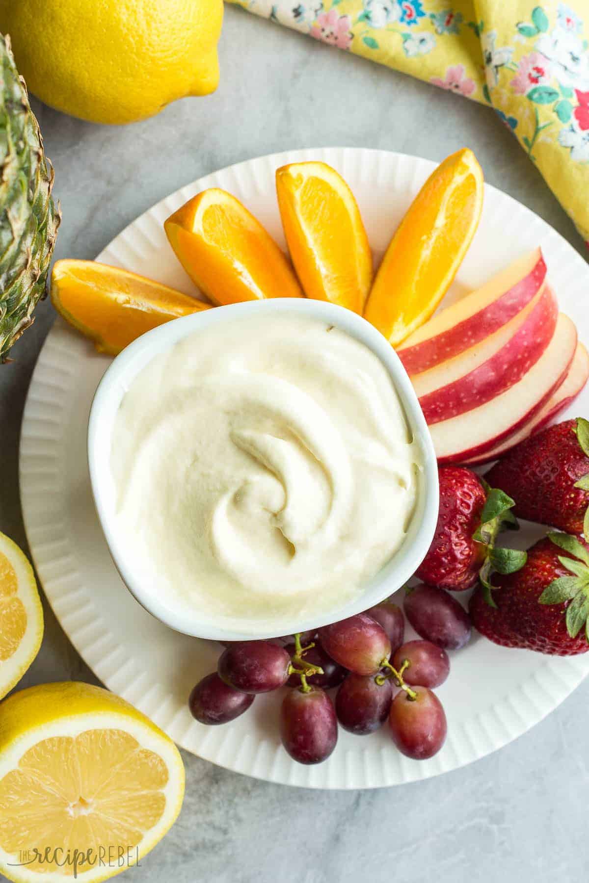 overhead image of pineapple lemonade fruit dip in white bowl with oranges apples strawberries and grapes