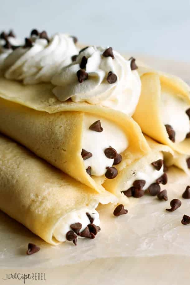 Soft homemade crepes filled with sweet ricotta cream and chocolate chips, topped with whipped cream and more chocolate chips. A breakfast version of an Italian favorite! close up image of cannoli crepes topped with whipped cream swirls with mini chocolate chips