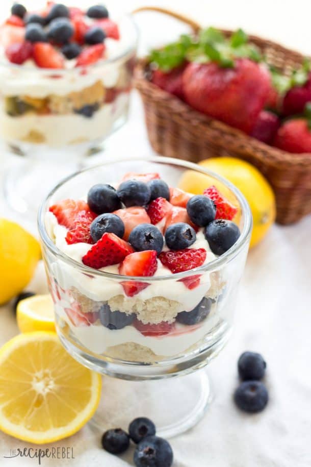 close up image of lemon berry cheesecake trifle in glass bowl with lemons and berries around
