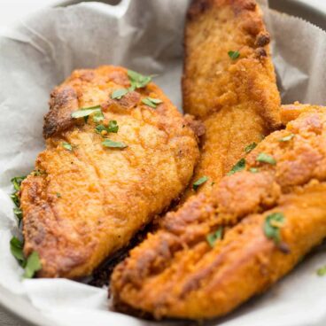 Three pieces of the best oven-fried chicken breast in a gray serving dish lined with white paper. Chopped herbs garnish the chicken.