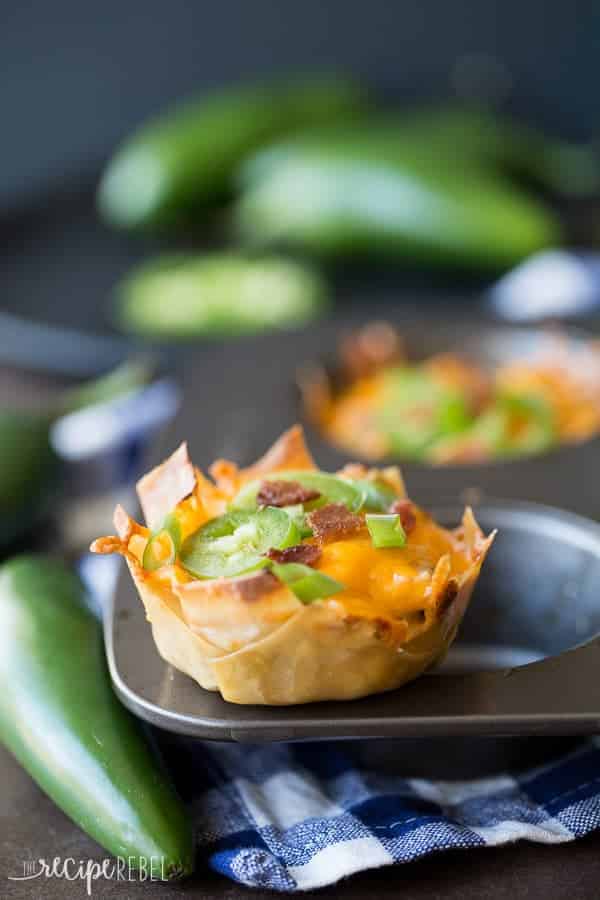 Creamy, spicy Jalapeno Poppers loaded with bacon, chicken and barbecue sauce, stuffed into a crispy wonton cup! jalalpeno popper wonton cups sitting on non stick muffin pan with whole jalapenos all around