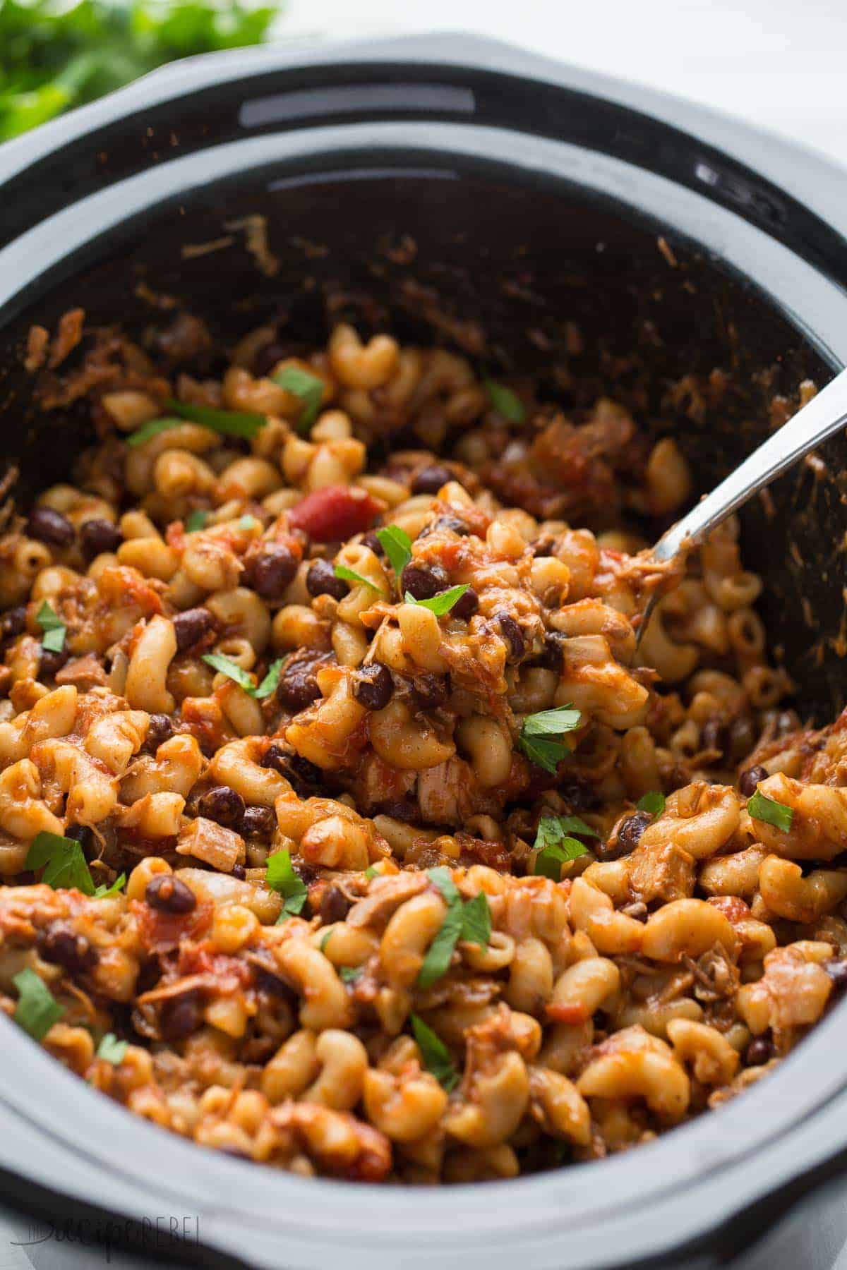 metal spoon scooping bbq chicken chili mac from black slow cooker