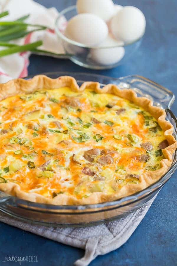 This Loaded Baked Potato Quiche is easy and full of flavor! It's the perfect easy quiche recipe for the holidays or a weeknight dinner! #breakfast #eggs #healthy #christmas #brunch #recipe easy loaded baked potato quiche in glass pie plate on blue background with fresh eggs in the background