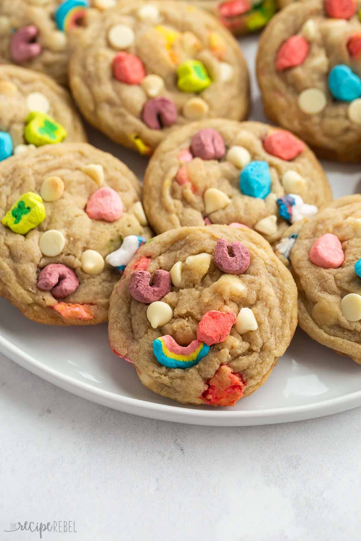 These White Chocolate Lucky Charms Cookies are perfectly chewy and made with Lucky Charms cereal and marshmallows. Perfect for St. Patrick's Day! Includes step by step recipe video. #cookies #recipe #dessert | st patrick's day | st. paddy's day | rainbow | cereal | cereal cookies | baking from scratch | cookie recipes close up image of lucky charms cookies on a white plate.