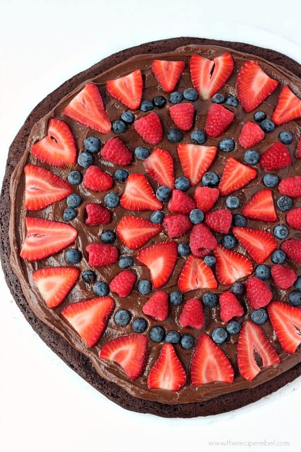 Nutella Brownie Fruit Pizza: a Nutella and cream cheese filling on a fudgy brownie base, topped with tons of fresh berries! Chocolate fruit pizza is the best kind! www.thereciperebel.com overhead image of round brownie topped with nutella and fresh berriess