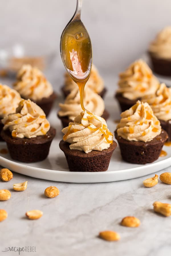 These Snickers Brownie Bites are made with perfectly fudgy mini brownies, topped with peanut butter frosting, caramel sauce and chopped salted peanuts. A sweet and salty treat! #brownies #chocolate #peanutbutter #dessert #dessertrecipe snickers brownie bites being drizzled with caramel on white plate