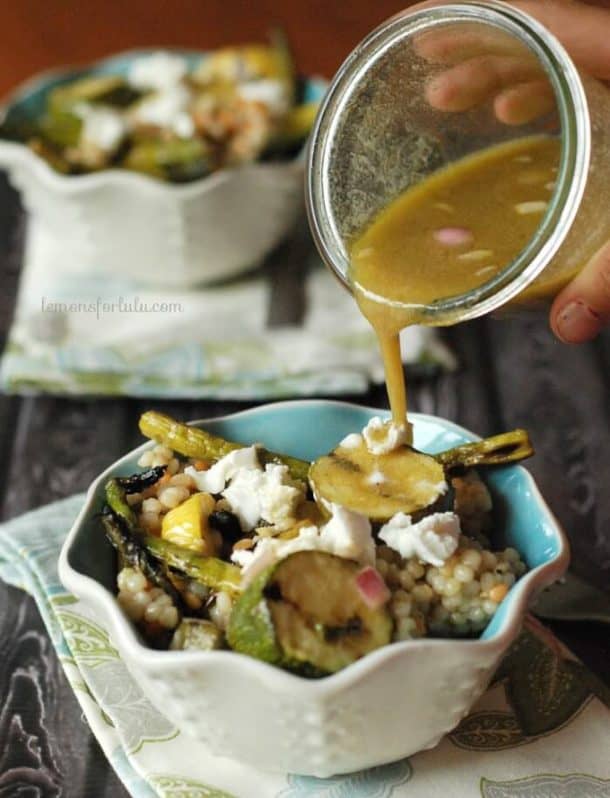 grilled veggies couscous salad in white and blue bowl with dressing being poured on