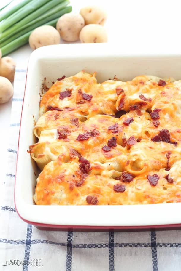 Perogy Stuffed Shells: garlic herb mashed potatoes and cheddar cheese stuffed inside a large pasta shell, and topped with creamy Béchamel sauce, more cheddar cheese and bacon before baking. An indulgent, make-ahead side dish that’s perfect for leftover mashed potatoes! www.thereciperebel.com red and white baking dish filled with perogy stuffed shells topped with bacon