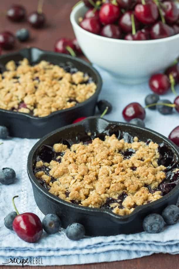 berry cherry crisp in individual cast iron skillets on blue towel with cherries and blueberries all around