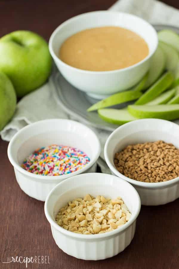 caramel apple dip in white bowl with green apple slices and sprinkles peanuts and toffee bits on the side