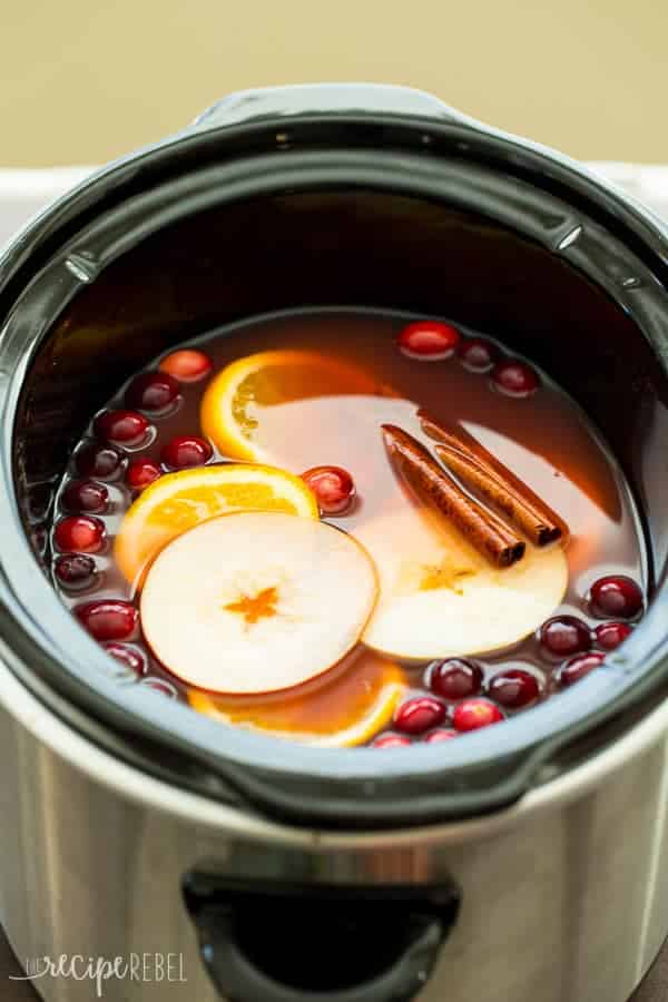 slow cooker cranberry apple cider in black crockpot with apple slices orange slices cinnamon sticks and cranberries