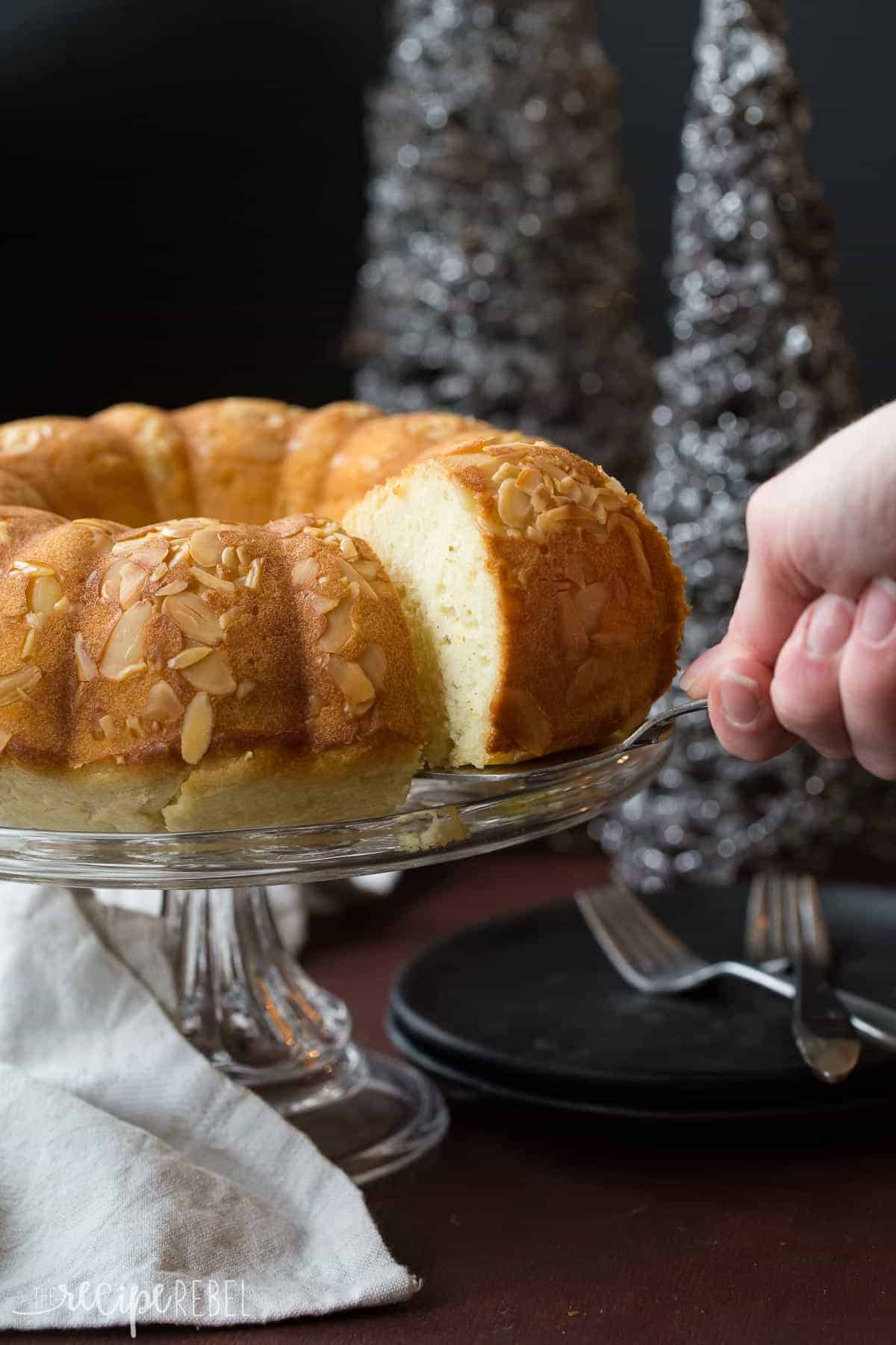 slice of almond pound cake being pulled out of the whole on the glass cake plate