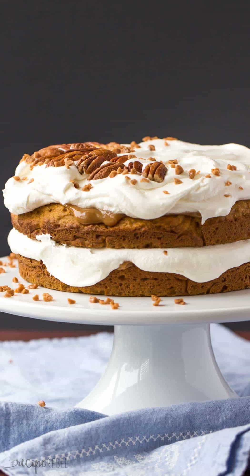 two layer pumpkin cake topped with whipped cream caramel sauce and pecans on white cake plate