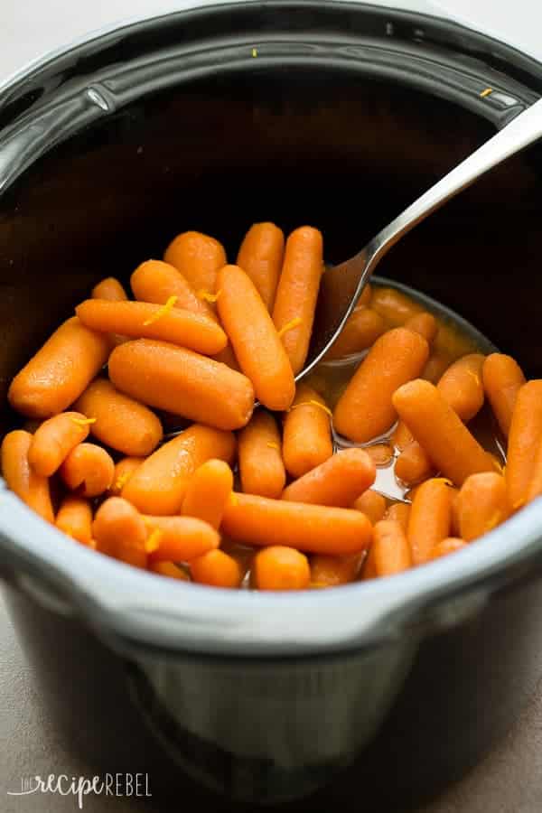 whole baby carrots in black slow cooker with glaze and metal spoon scooping some