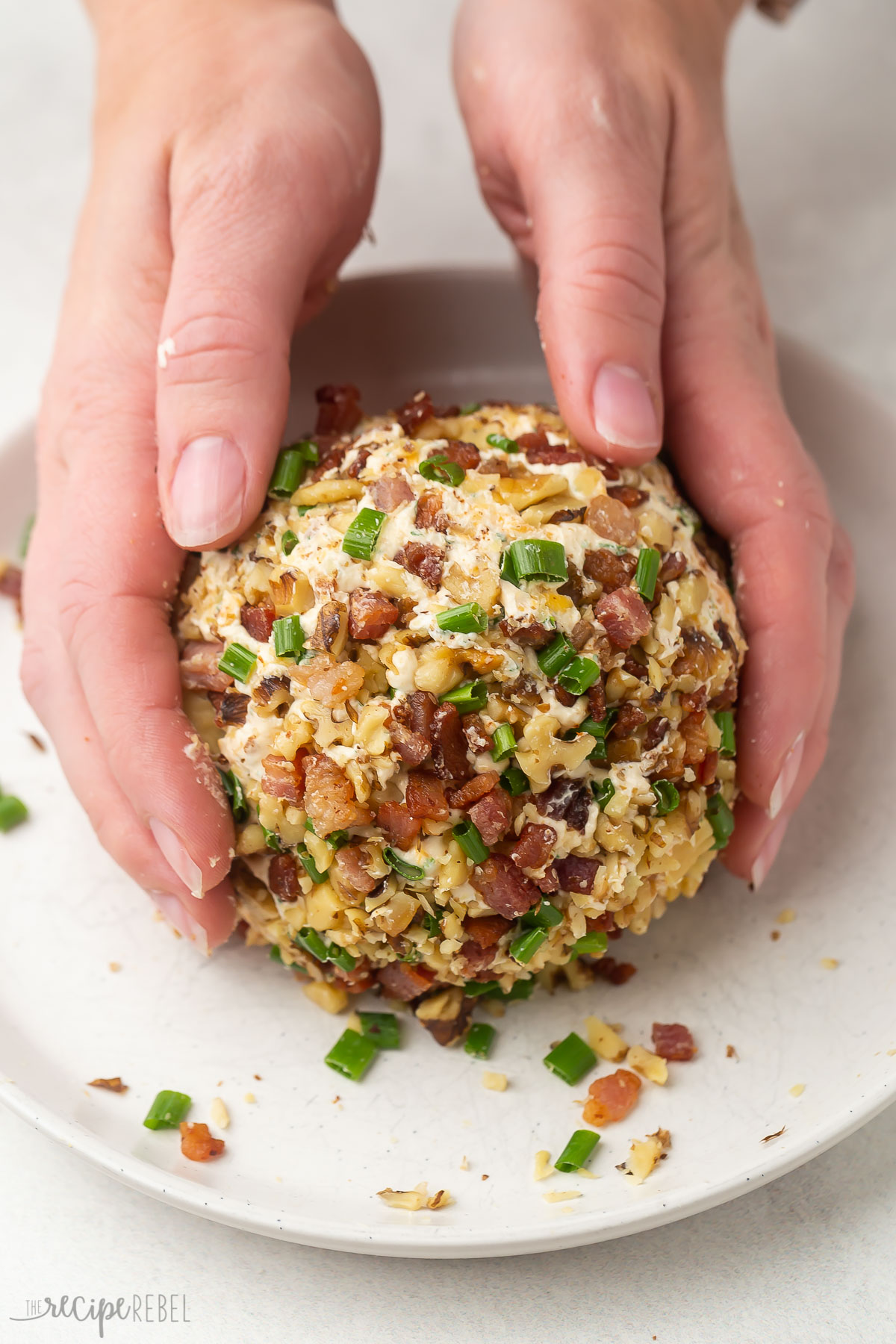 two hands covering the cheese ball in bacon nuts and green onions.
