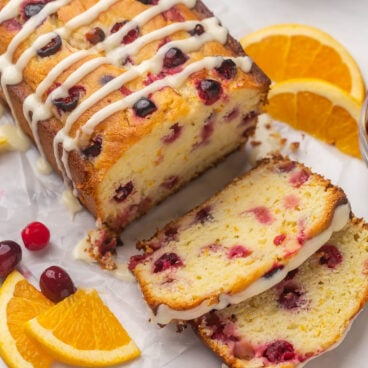 side angle of whole cranberry orange bread with two slices cut and orange slices and cranberries on the side.