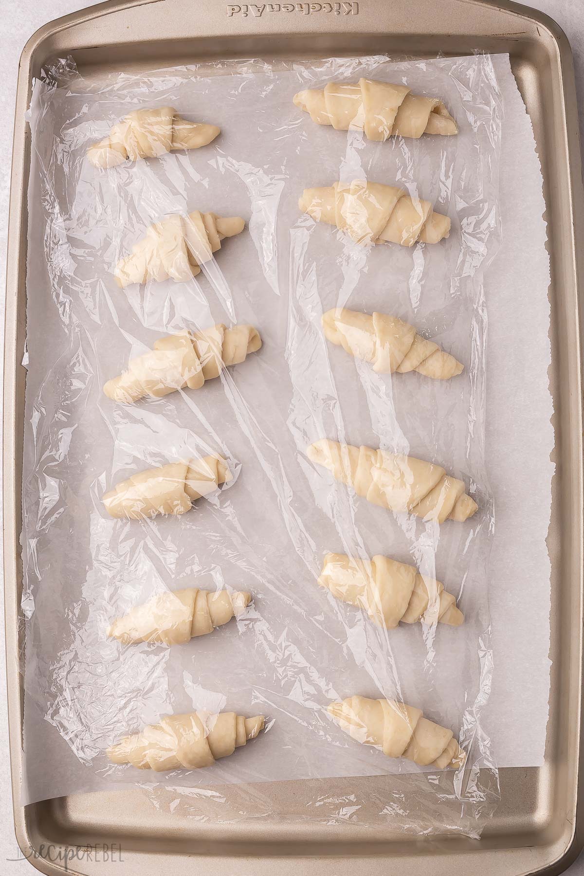 rolled crescents on baking sheet with parchment paper covered with plastic wrap to rise.