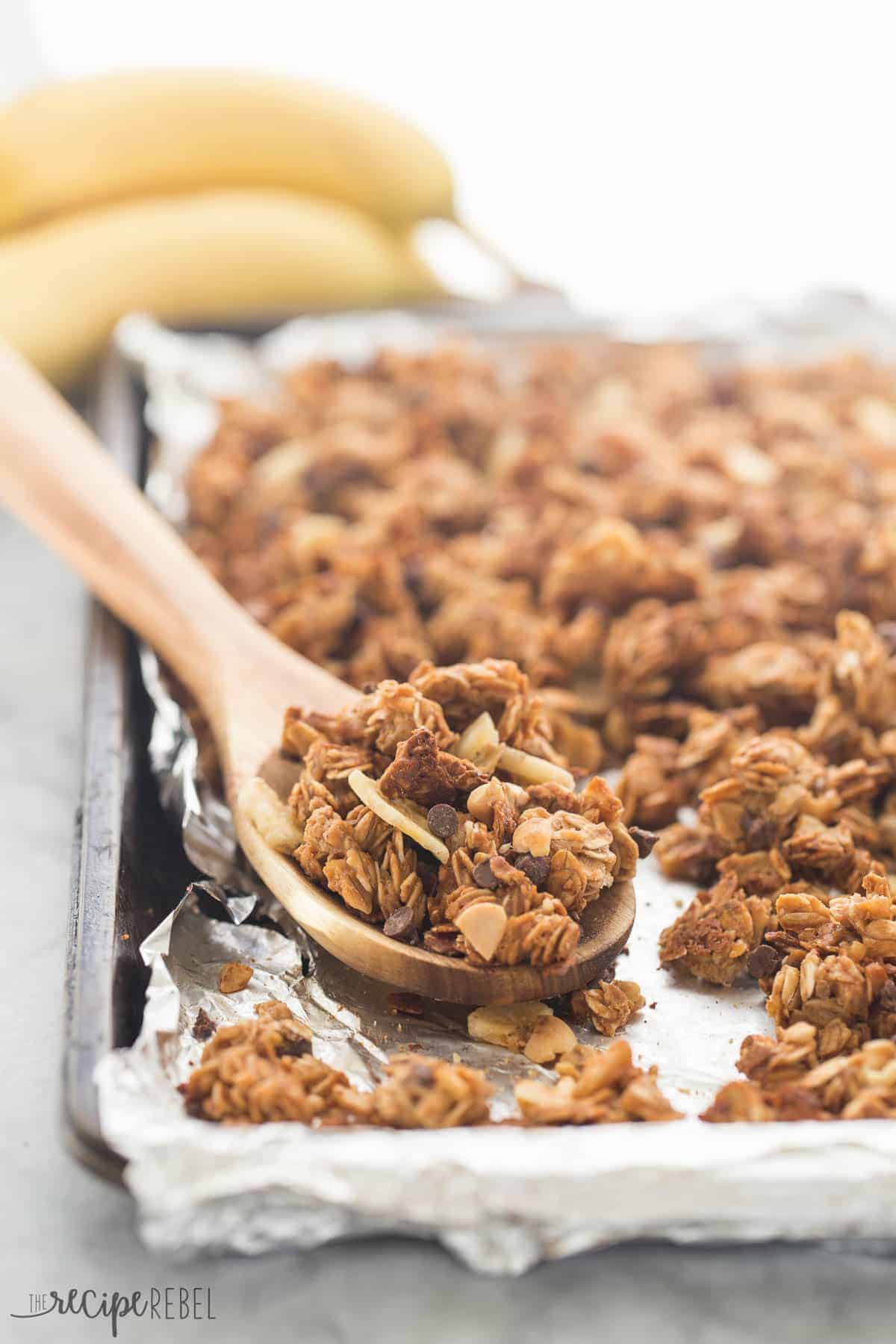 chunky monkey granola baked on a sheet pan lined with foil and some in a wooden spoon