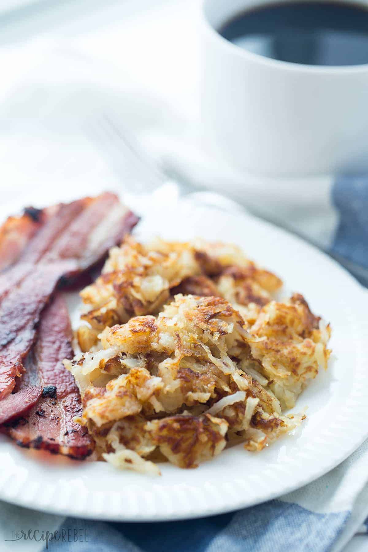 pile of crispy hashbrowns on white plate with two pieces of bacon and blue towel underneath