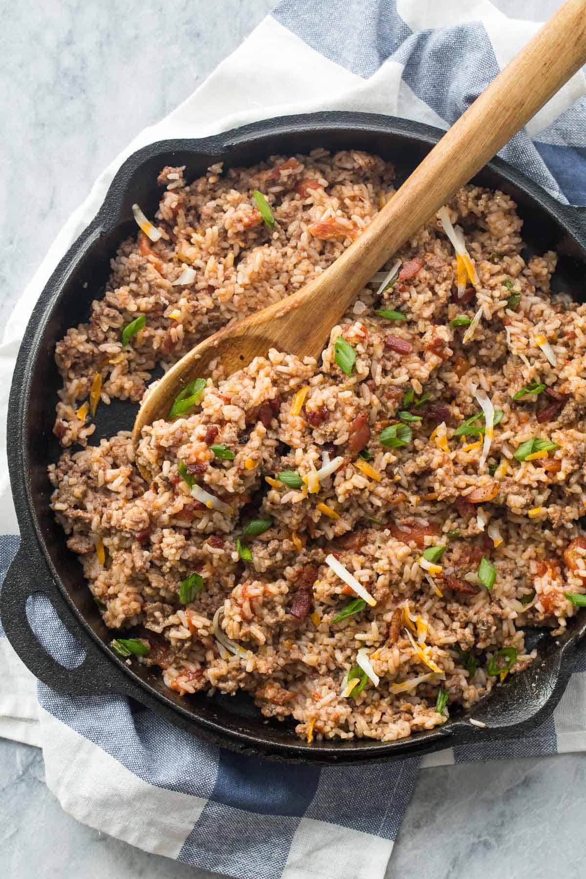 cast iron skillet overhead with bacon cheeseburger rice in it scooping with wooden spoon