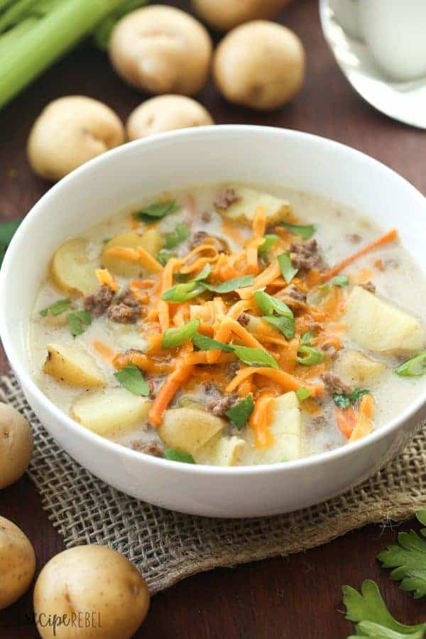 cheeseburger crockpot soup in white bowl with shredded cheddar cheese on top and potatoes in the background