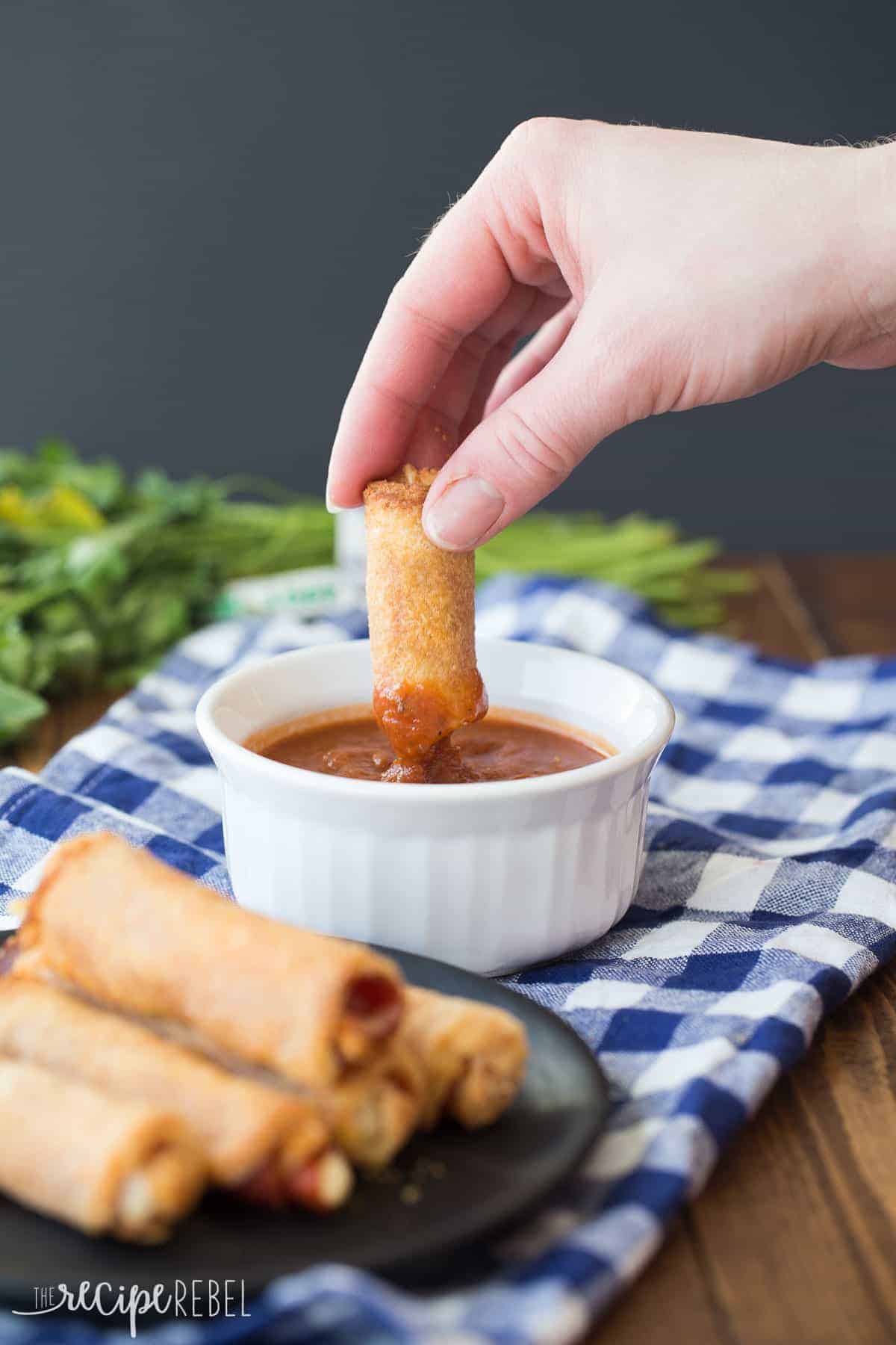 hand dunking pizza roll up in small white bowl of pizza sauce over checkered napkin
