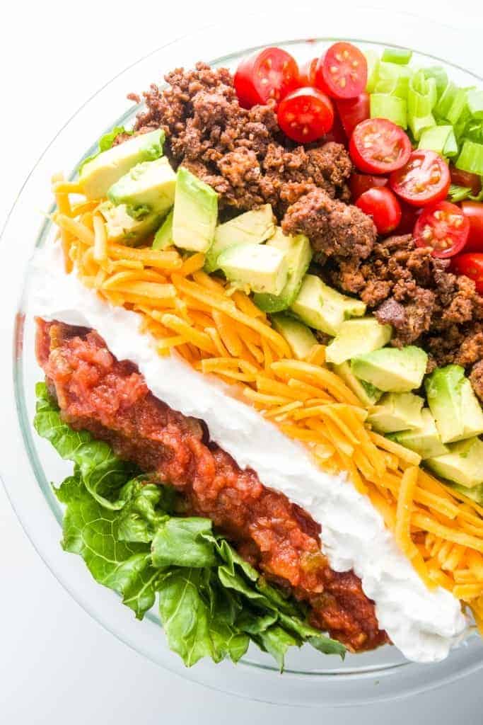 healthy taco salad in large clear bowl with layers of cheese avocado beef tomatoes and green onions