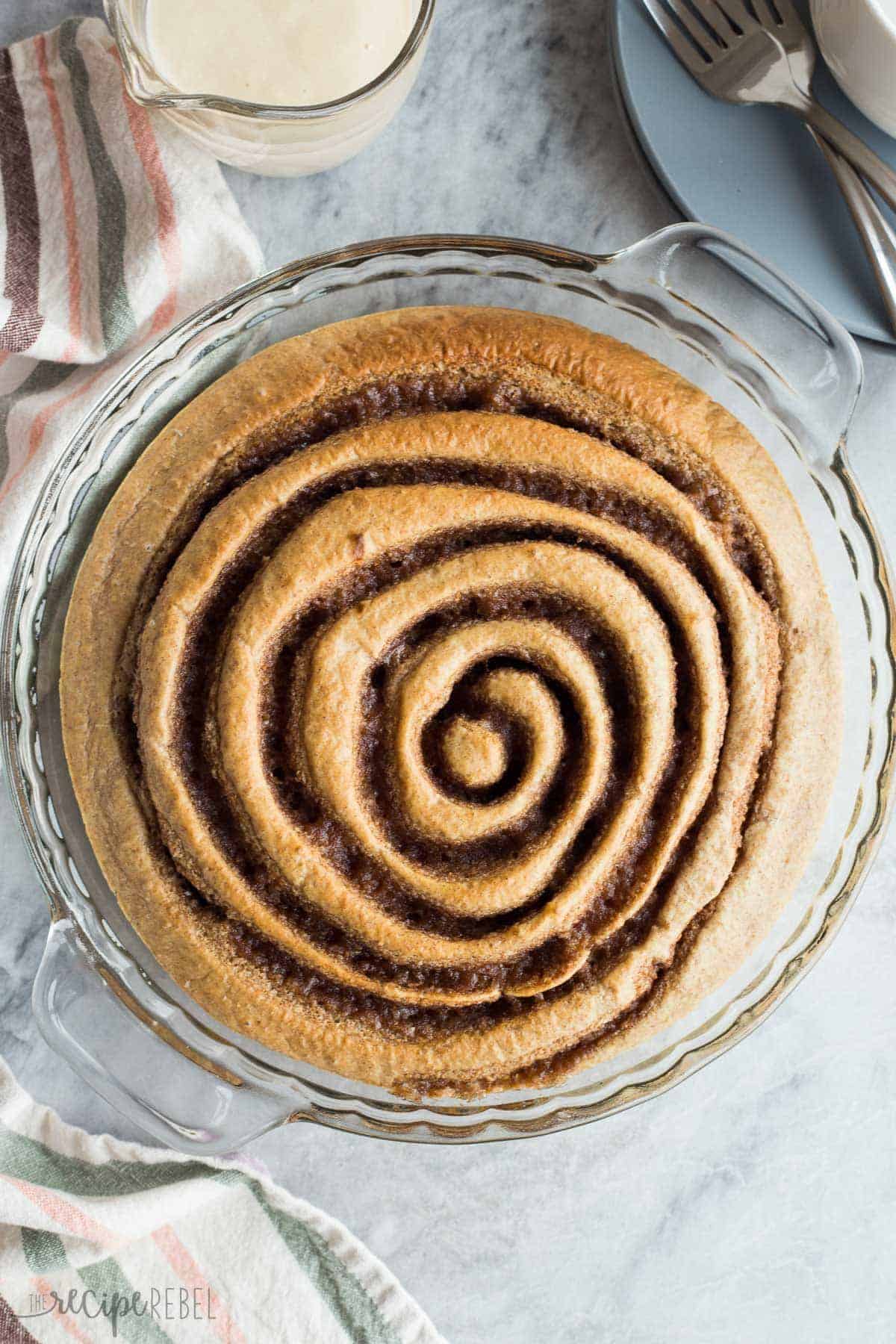 This Healthier Cinnamon Roll Baked Pancake is whole wheat, low in sugar, and bakes in a pie plate so there's no flipping or waiting! Drizzle with easy Maple Cream Cheese Syrup for a special treat! overhead image of healthier cinnamon roll baked pancake in glass pie plate on marble
