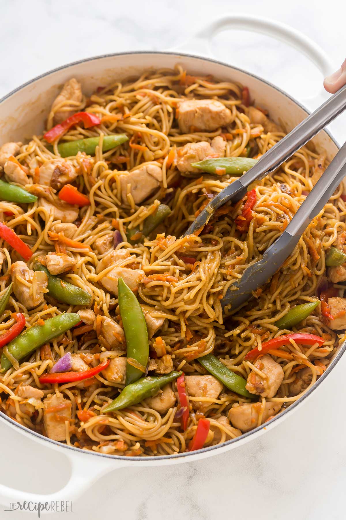 close up image of tongs grabbing chicken chow mein noodles in pan