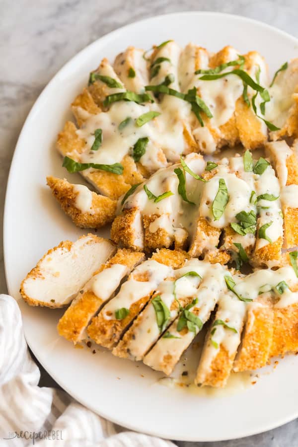sliced crispy chicken breasts on white platter drizzled with creamy sauce and fresh basil