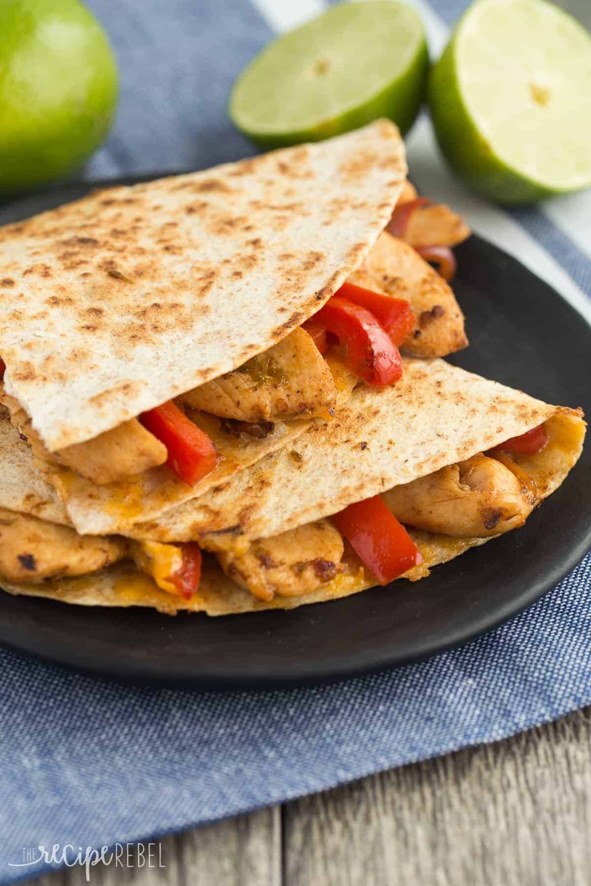 Honey Lime Chicken Quesadillas are filled with chicken, peppers, onions and cheese and are the perfect easy weeknight or summer meal! My whole family LOVES these! honey lime chicken quesadillas on a black plate with red peppers and lime halves in the background