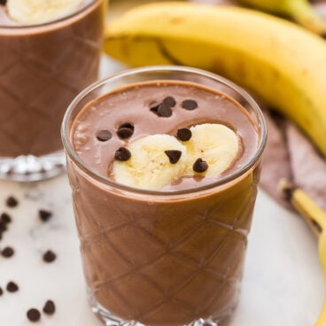 two glasses of chocolate peanut butter smoothie with bananas in the background