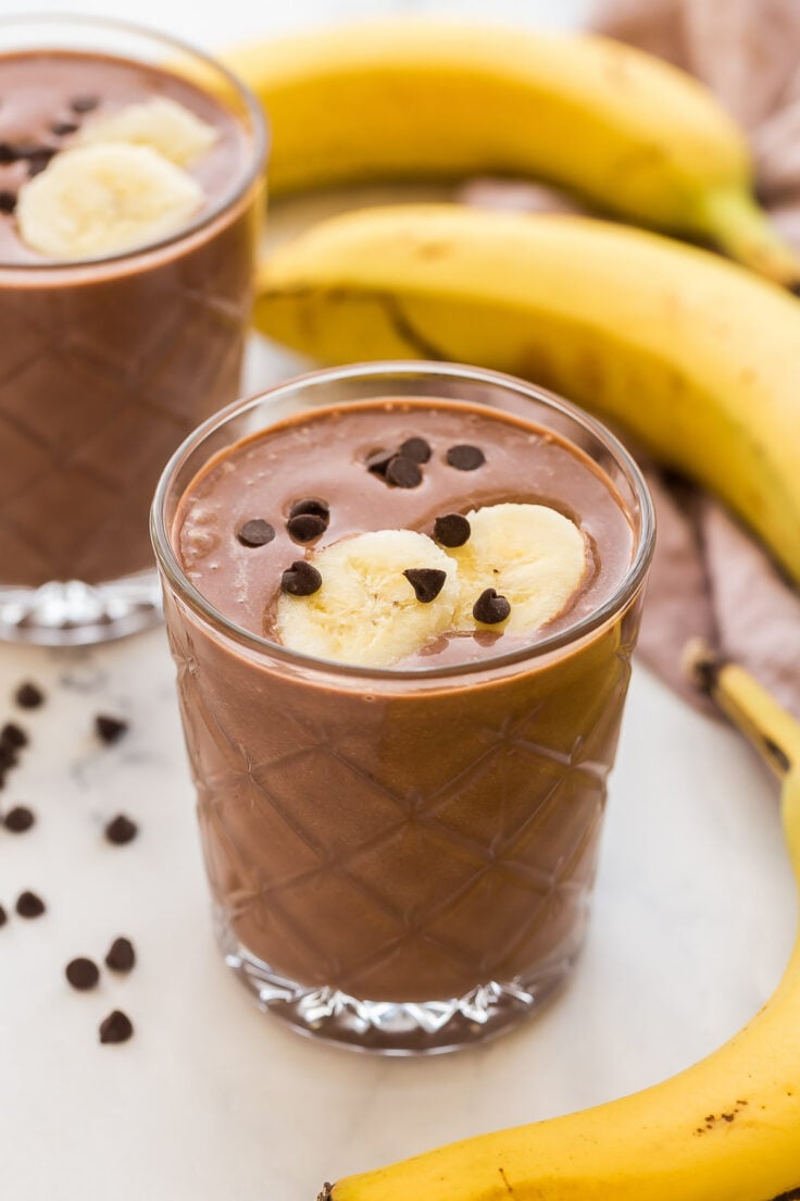 two glasses of chocolate peanut butter smoothie with bananas in the background