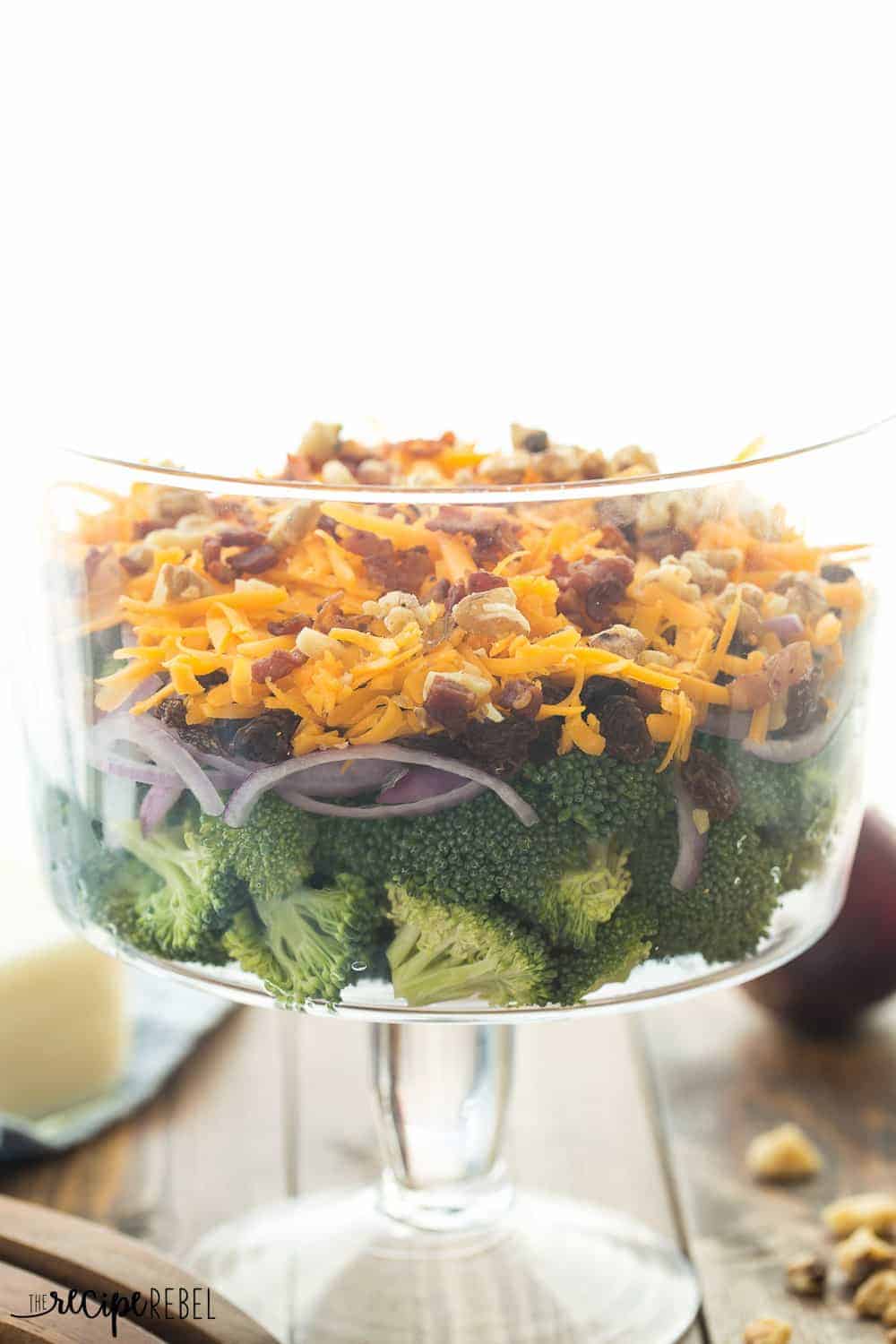 glass trifle bowl with layers of broccoli salad including red onion cheddar cheese nuts and raisins