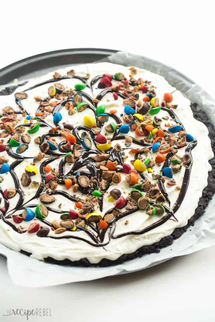 This Frozen Ice Cream Dessert Pizza is THE no bake summer dessert you need! A Dairy Queen Copycat Treatzza Pizza, it has an Oreo fudge crust topped with ice cream, fudge sauce and M&M's! https://www.thereciperebel.com/frozen-ice-cream-dessert-pizza-treatzza-pizza/ frozen ice cream dessert pizza on pizza pan with chocolate sauce and M&Ms