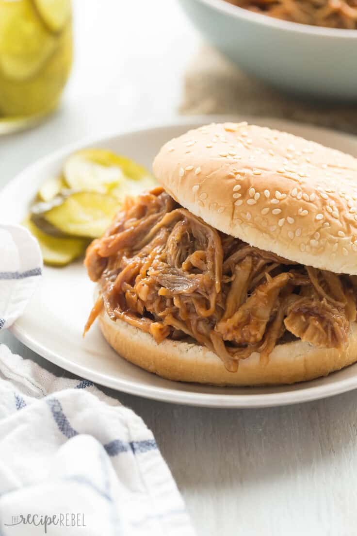 This easy Slow Cooker Pineapple Brown Sugar Pulled Pork recipe is smothered in homemade pineapple brown sugar barbecue sauce! Sweet, tangy and so tender! https://www.thereciperebel.com/slow-cooker-pineapple-brown-sugar-pulled-pork/ slow cooker pineapple brown sugar pulled pork on a bun on a plate with pickles on the side