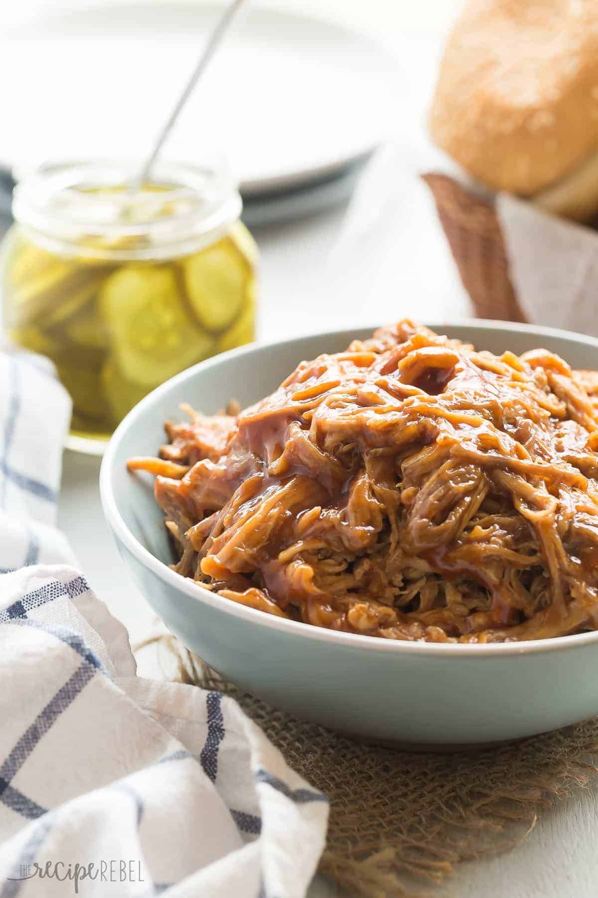 slow cooker pulled pork in blue bowl with pickles and buns in the background