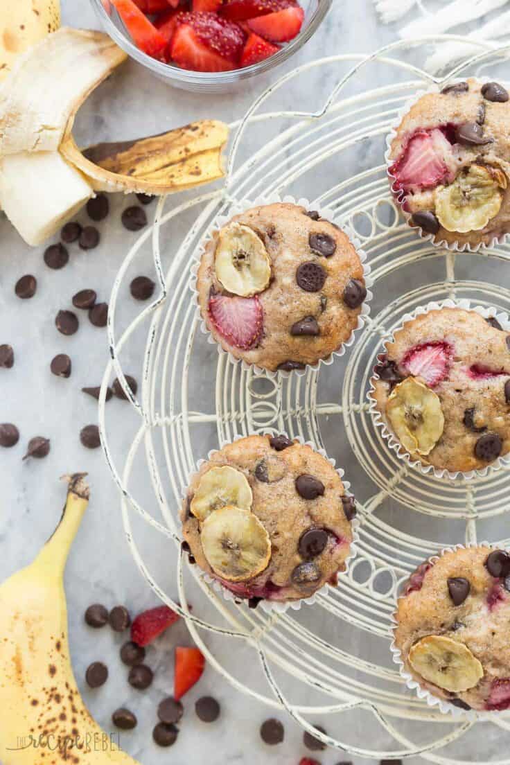 These Chocolate Chip Banana Muffins are the best -- so moist but made healthier with whole wheat flour, yogurt and loaded with fresh strawberries! strawberry banana chocolate chip muffins overhead on white cooling rack with bananas strawberries and chocolate chips all around