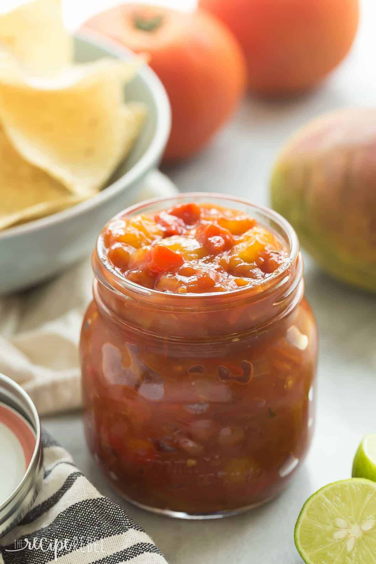 mango salsa in jar with lime wedges tortilla chips and mangoes in the background