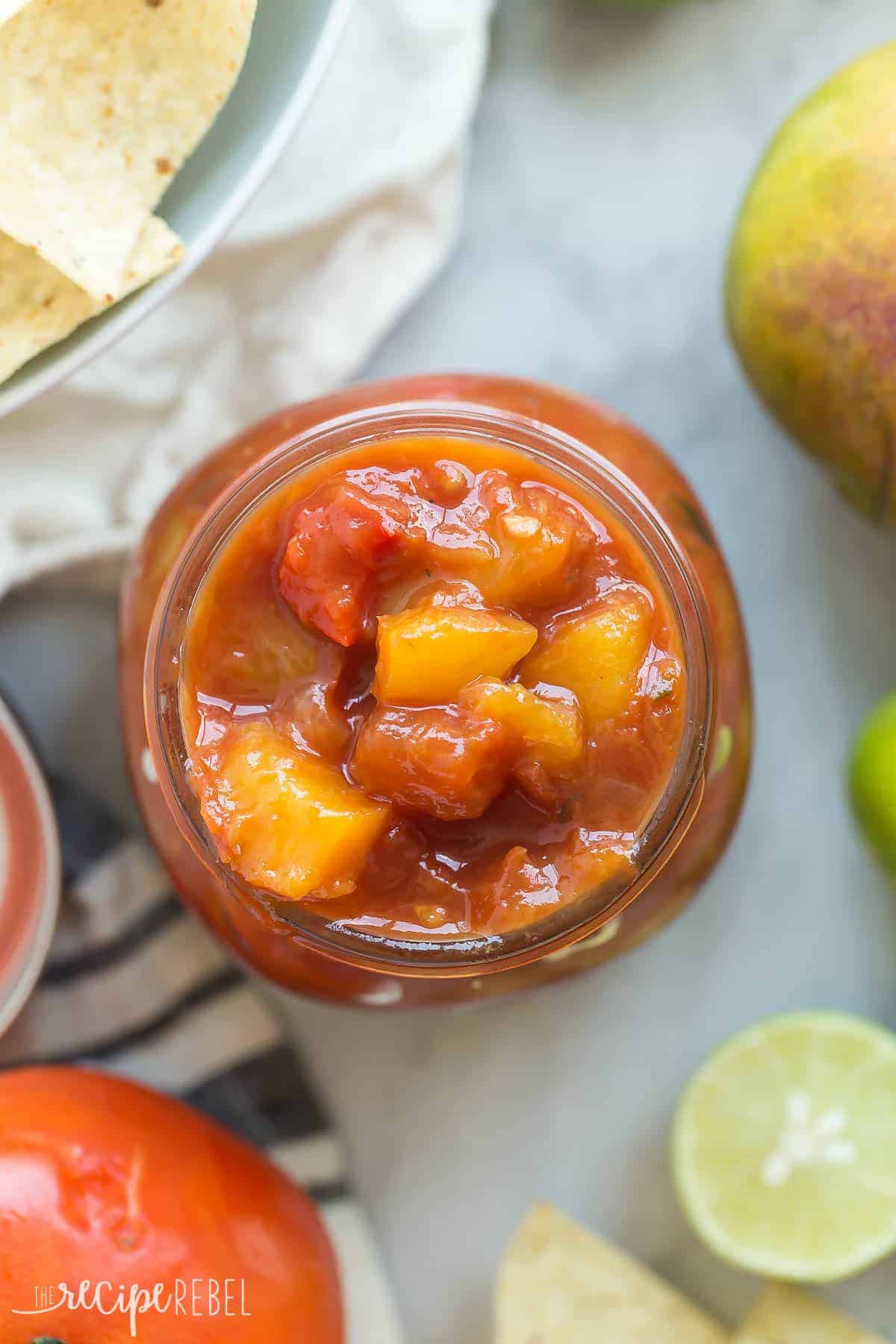 overhead image of mango salsa in jar with tomatoes and lime wedges all around