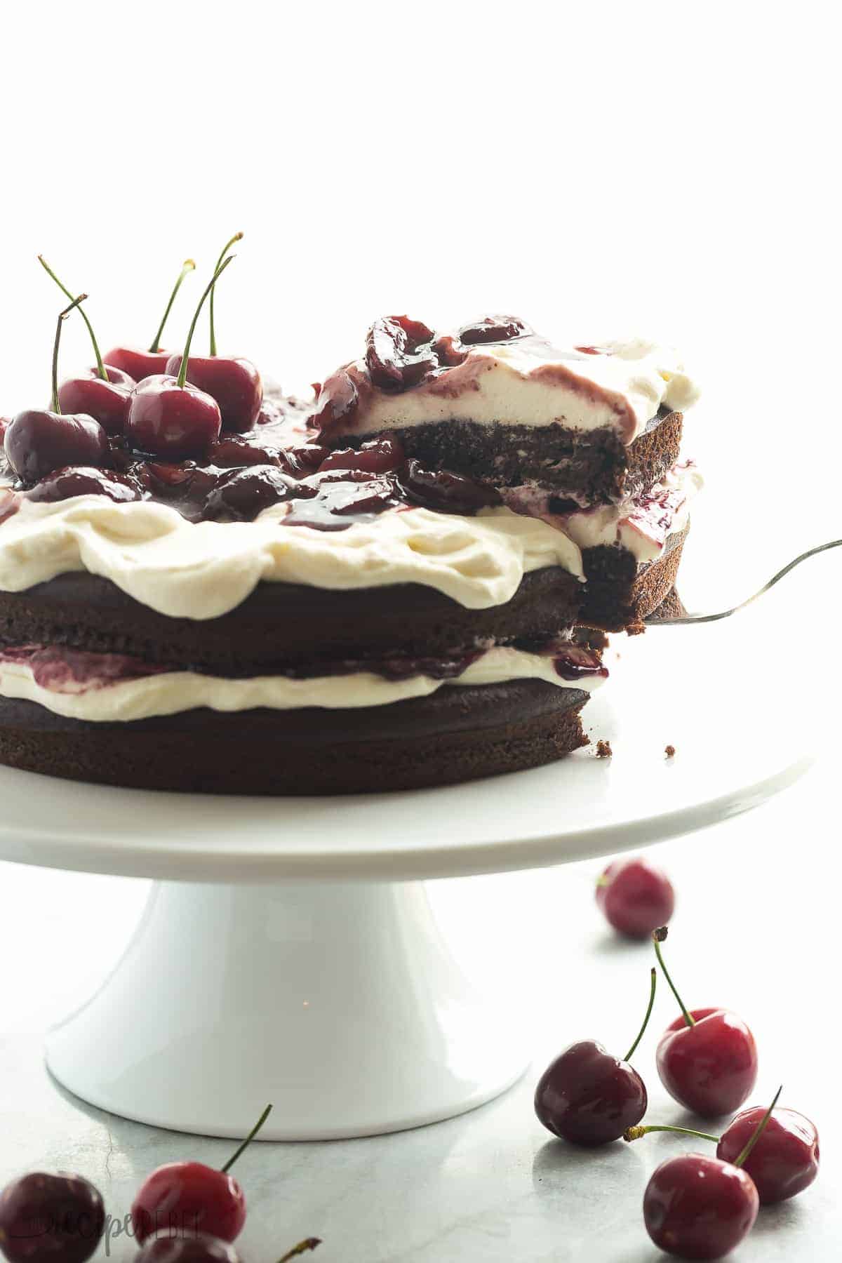black forest cake whole on white cake plate on white background with piece being removed