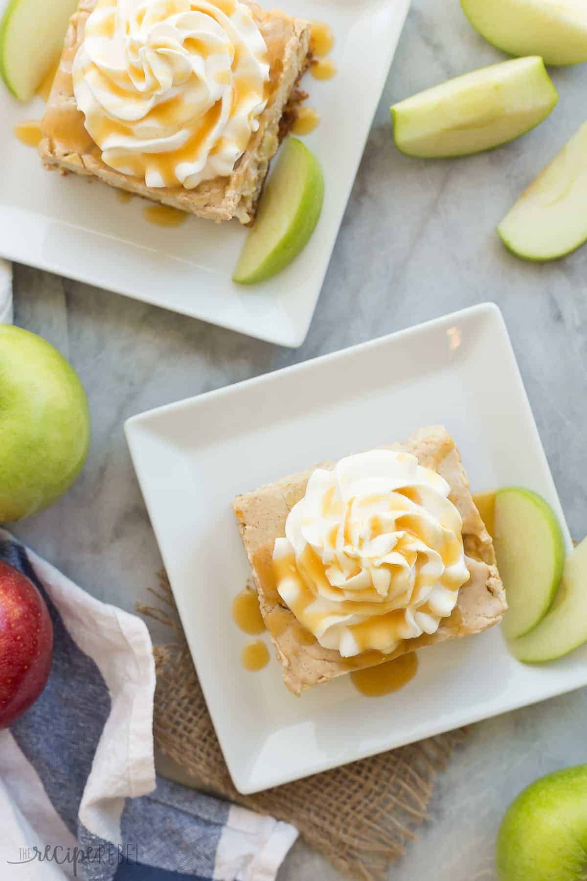 two pieces of caramel apple cheesecake bars on white plates with whipped cream and caramel sauce overhead