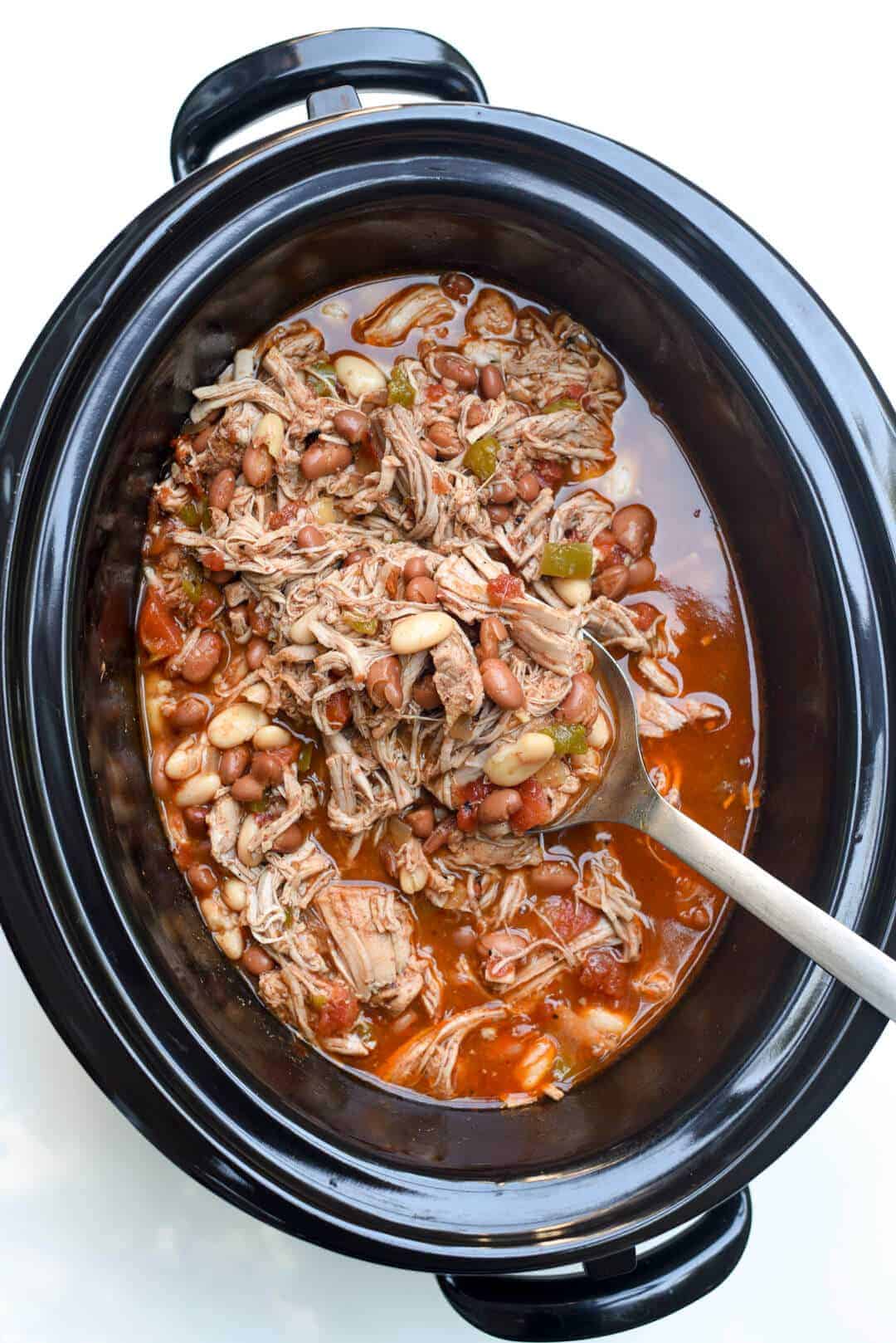 overhead image of black slow cooker with mexican pork and beans with metal spoon in pot