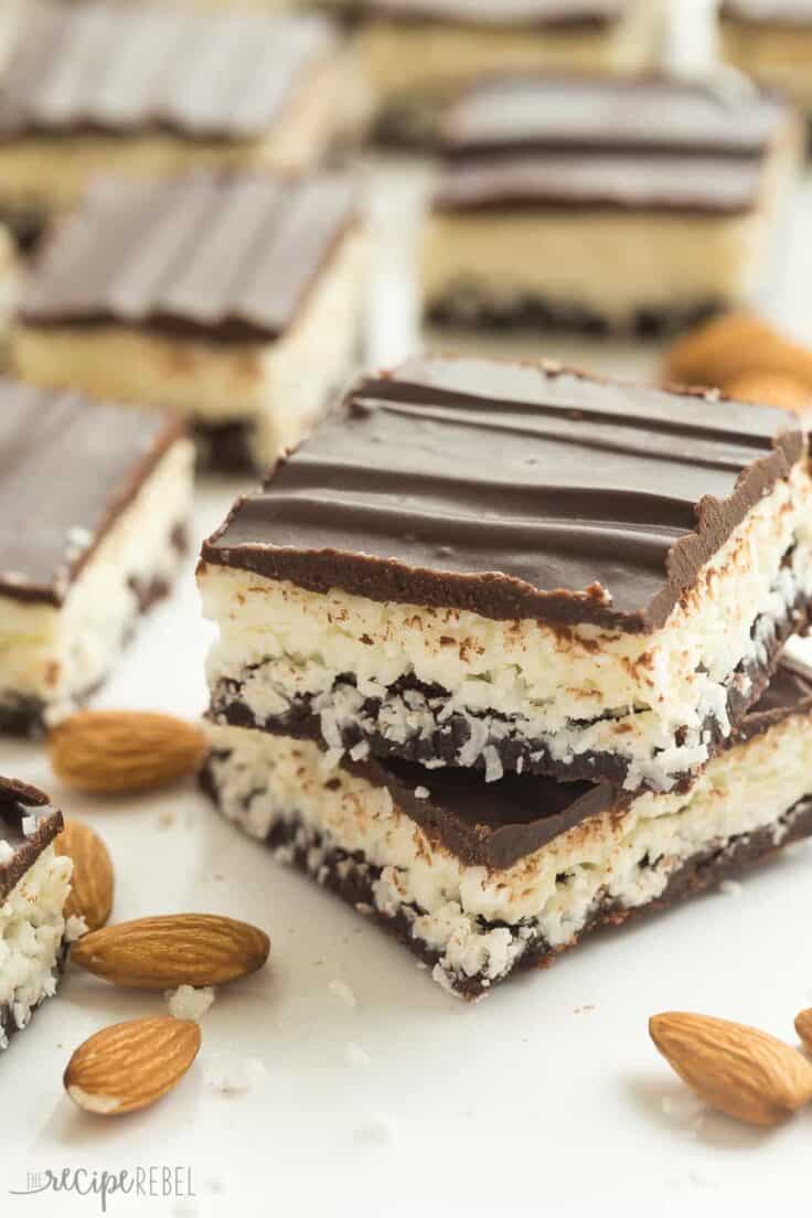 These Almond Joy Brownies have a thin, fudgy brownie layer and are topped with a creamy coconut filling, almonds and a layer of chocolate! They are an easy treat for the holidays! Two Almond Joy brownies stacked on top of each other with scattered almonds and more brownies in the background.