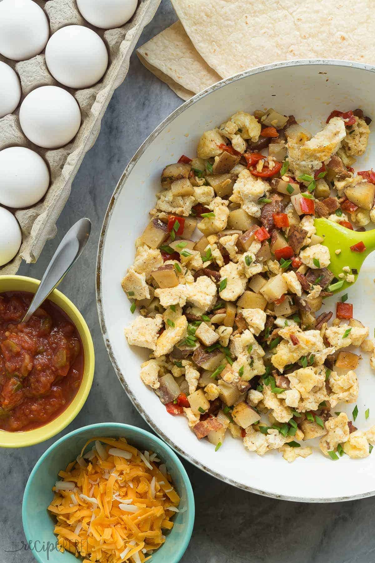 These are our FAVORITE Breakfast Burritos! They are loaded with homemade hashbrowns, peppers, bacon, eggs, salsa and cheese! They are make ahead and freezer friendly -- perfect for busy weeknights. overhead image of components of breakfast burritos including scrambled eggs tortillas cheese and salsa