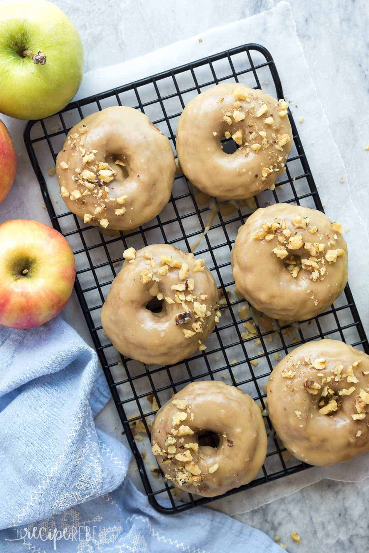 6 apple cinnamon baked donuts on black cooling rack on marble background with whole apples on the side