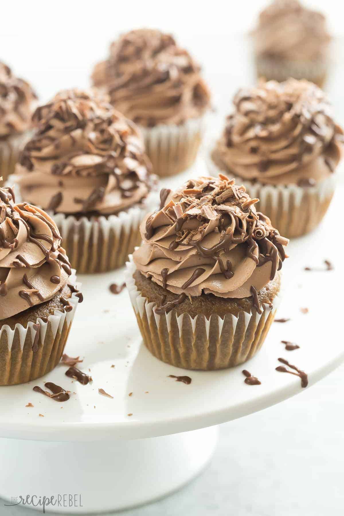 These Mocha Nutella Cupcakes are the perfect way to get your coffee fix! A moist coffee flavored cupcake topped with creamy Nutella frosting! white cake plate full of mocha nutella cupcakes with frosting and chocolate drizzle