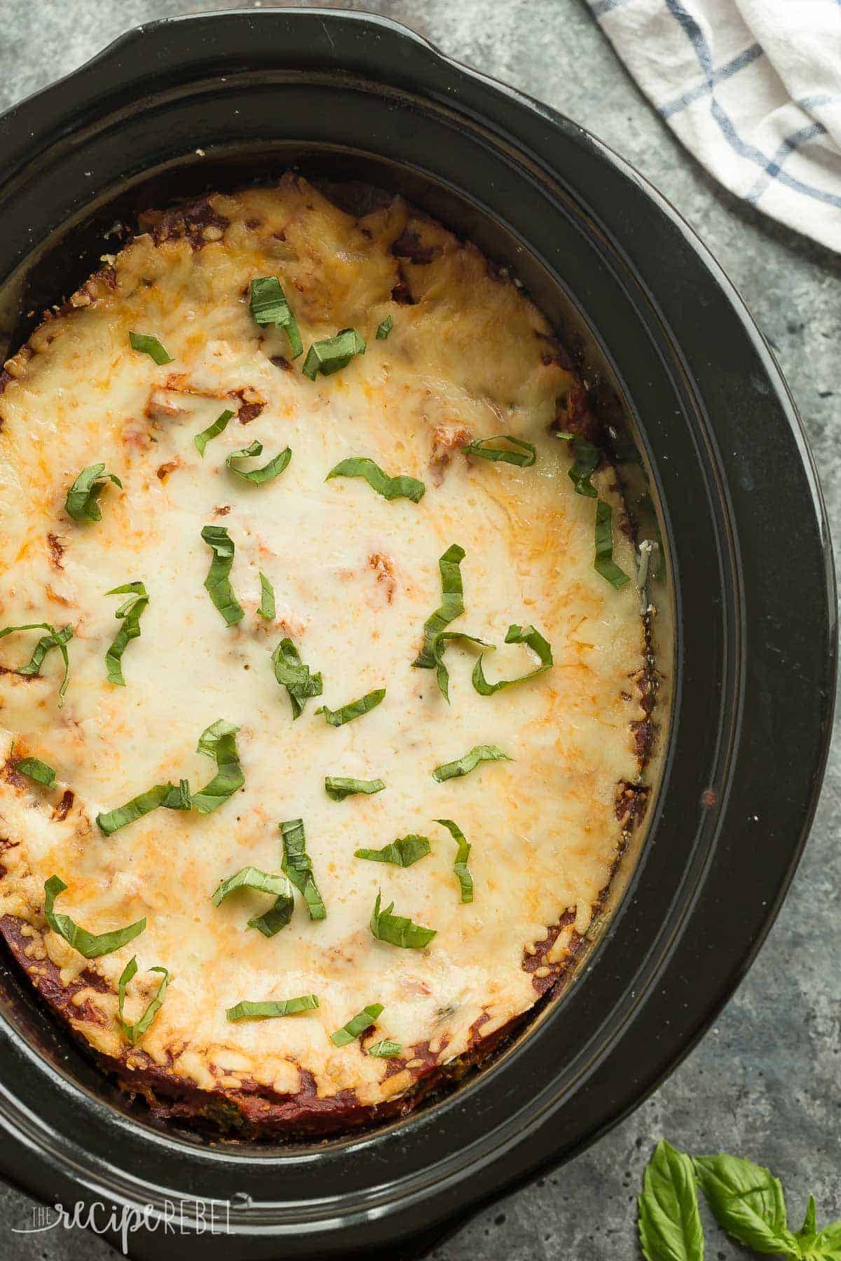 overhead image of black slow cooker with whole slow cooker lasagna and melted cheese on top