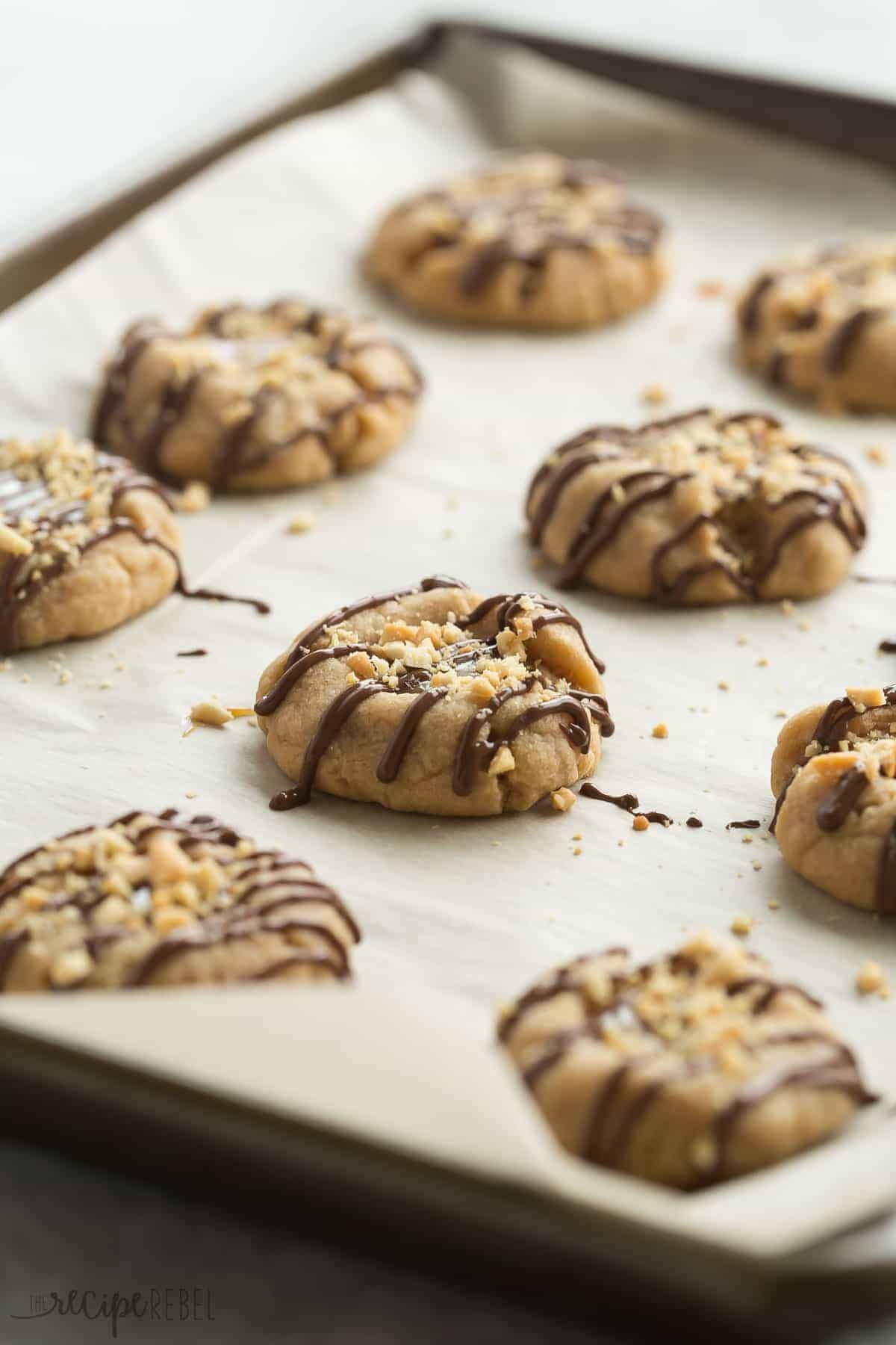 These Peanut Butter Turtle Thumbprint Cookies are a new twist on a classic Christmas cookie -- with a peanut butter cookie base, a simple caramel filling, a drizzle of chocolate and a sprinkling of peanuts, they're sure to be a new holiday tradition! peanut butter turtle thumbprint cookies on a parchment lined sheet pan with chocolate drizzle